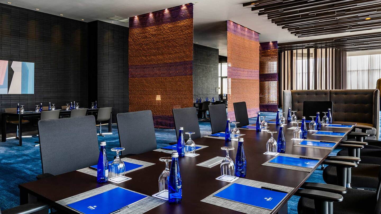 Salón interior con los pisos cubiertos por un tapete azul, del techo cuelgan unos tapices. Hay una mesa larga rectangular color café, sobre la que hay varias botellas de agua y copas. Varias sillas rodean la mesa.