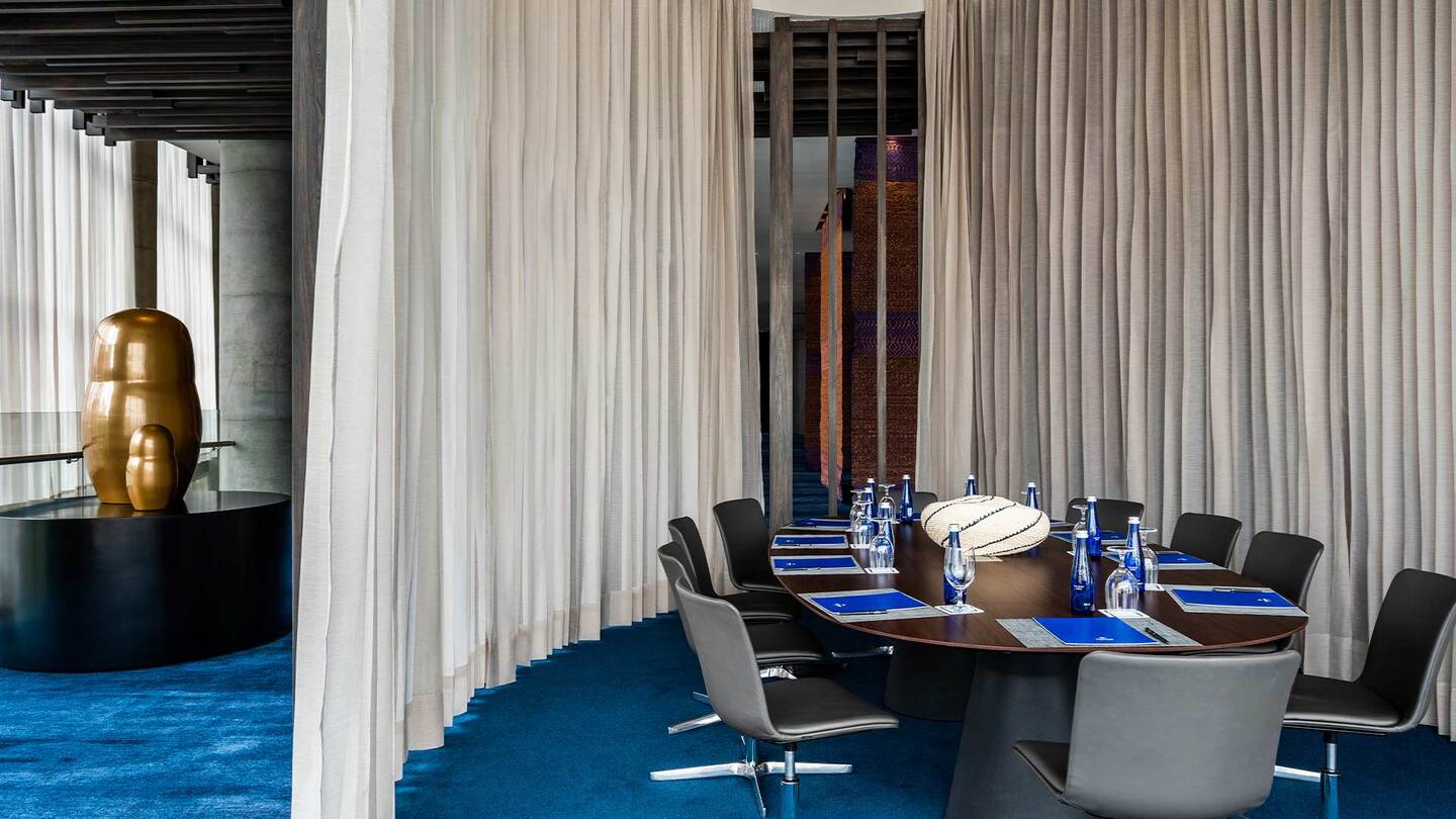 Salón en donde a través de una manta que cuelga del techo se divide una mesa ovalada y rodeada de varias sillas. En la mesa hay varias botellas de agua, copas de cristal y una especia de cuadernos con esferos.