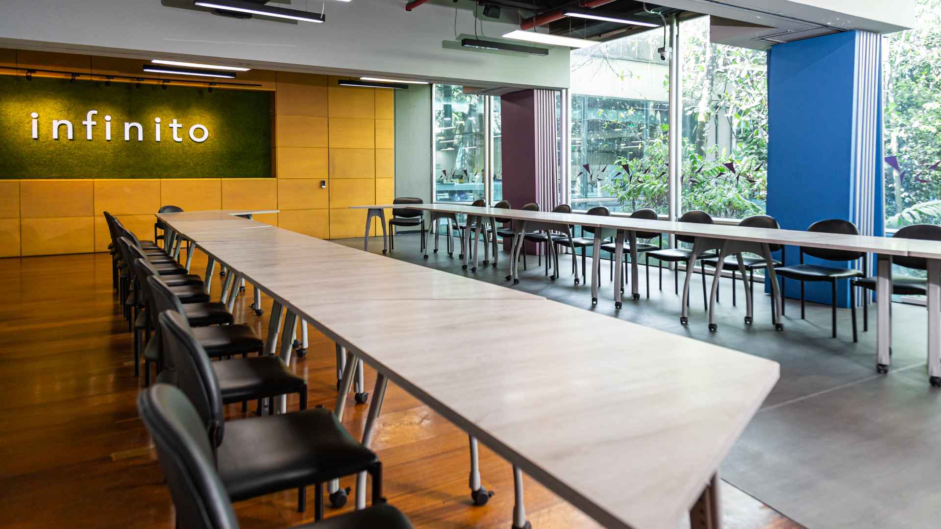 Salón amplio con un par de mesas rectangulares, largas y varias sillas alrededor. Hay una pared en madera y una ventana grande hacia una zona de árboles.