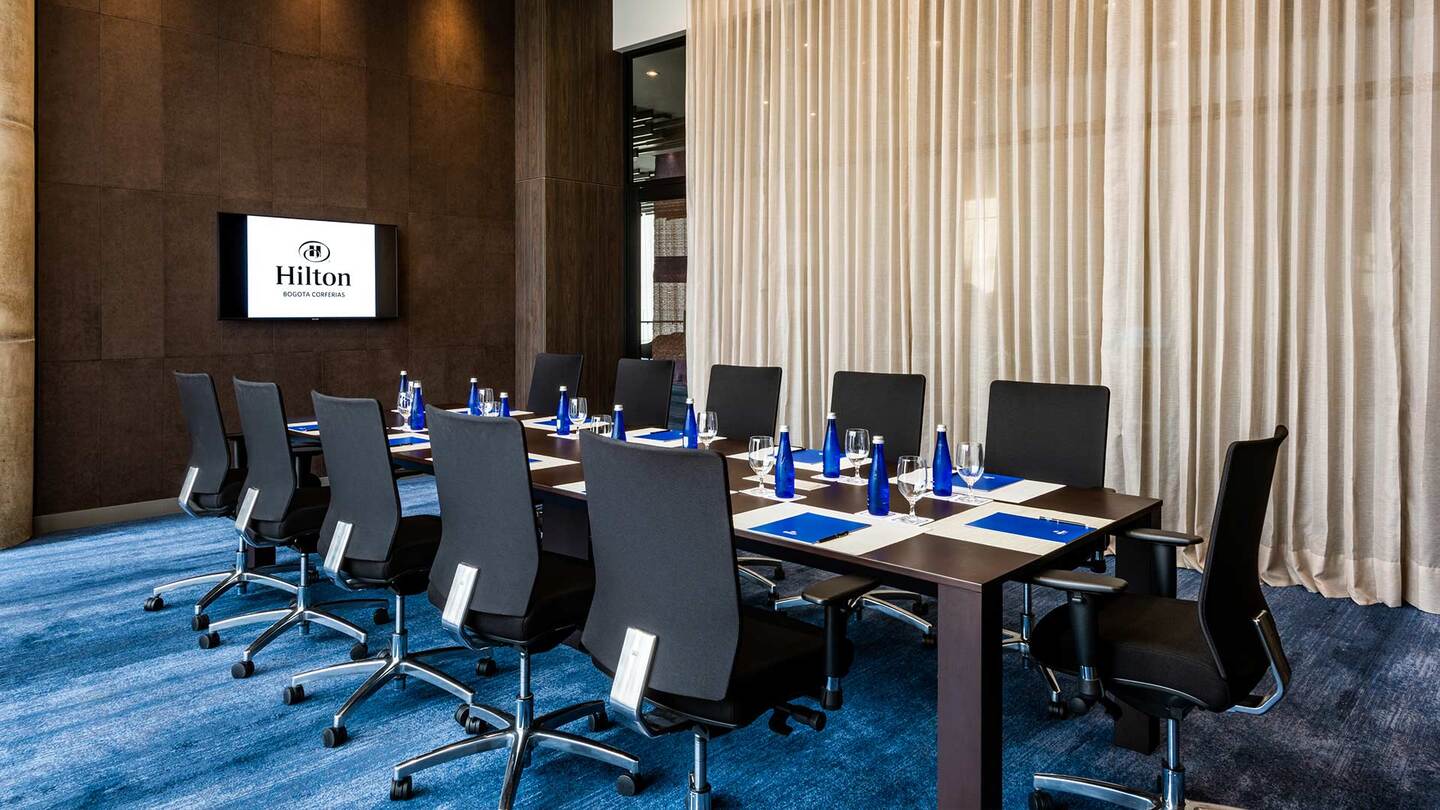 Salón interior con los pisos cubiertos por un tapete azul, del techo cuelgan unos tapices. Hay una mesa larga rectangular color café, sobre la que hay varias botellas de agua y copas de cristal. Varias sillas rodean la mesa. A un lado hay un ventanal cubierto por una larga cortina blanca. Al frente hay una pared en la que sobresale un letrero que dive "Hilton".