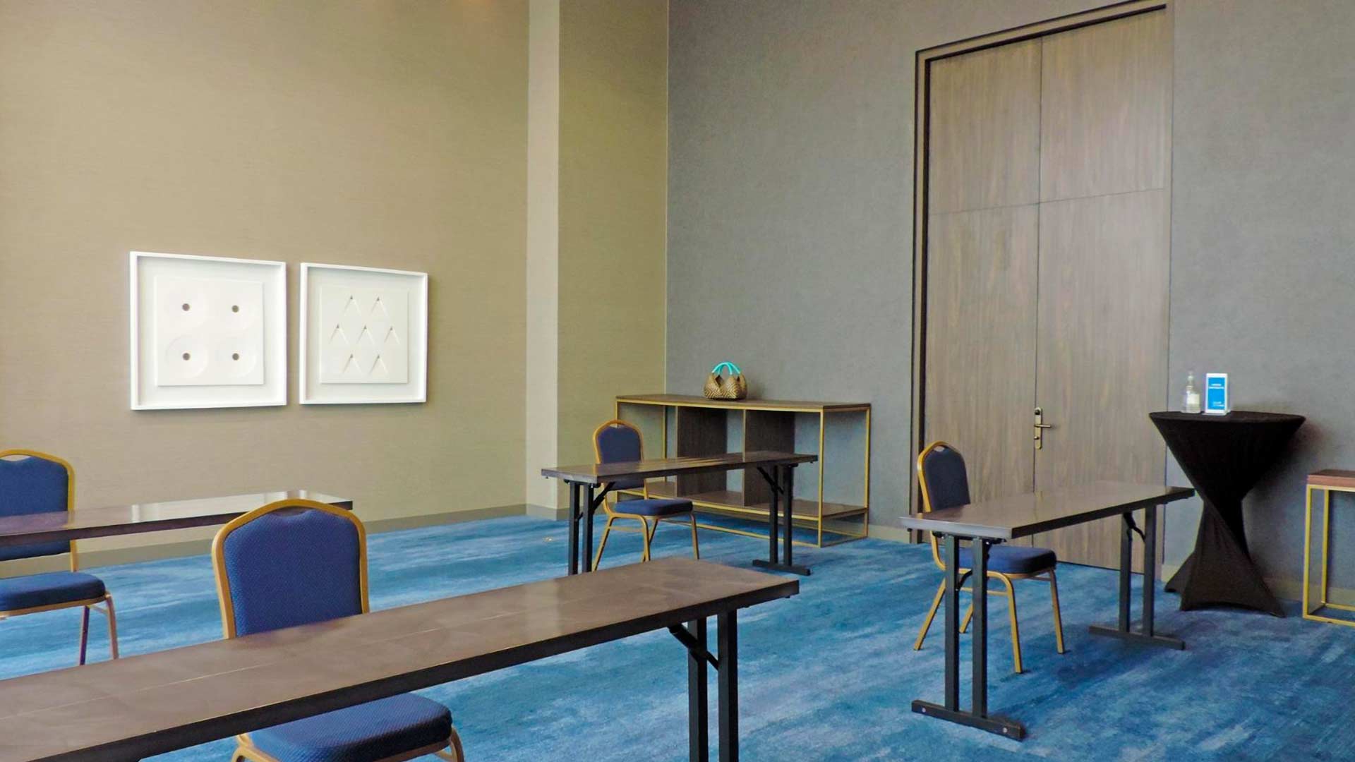 Salón con cuatro cortas mesas rectangulares, cada una con una silla sencilla. Hay una pared de color marrón claro. El piso está cubierto por un tapete azul. A un lado hay una puerta de madera y está cerrada.