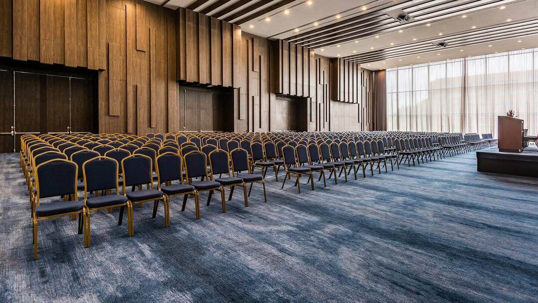Salón grande con un centenar de sillas y un atril al frente de ellas. Las paredes están ciubiertas de madera, el piso de un tapete azul y hay un ventanal grande a un costado, por donde entra muy buena luz natural.