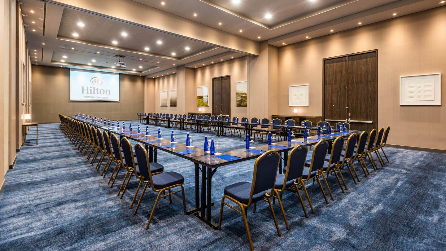 Salón organizado con una fila de mesas formando una U. Las paredes son color marrón claro. hay dos puestas de acceso que están cerradas. Hay varios bombillos de luz en el techo y un videobeam. Hay un tapete grande de color azul.