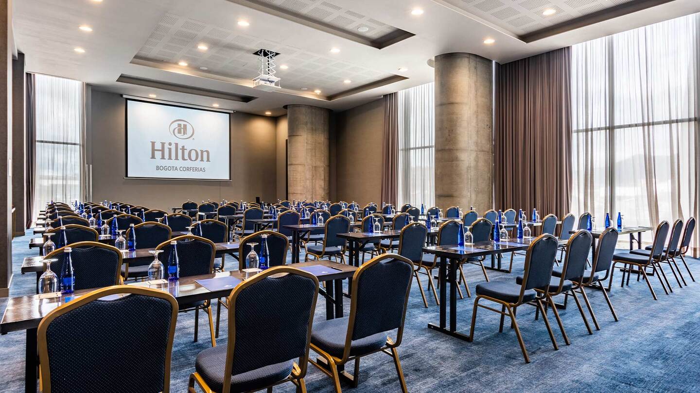 Salón con varias mesas rectangulares y sillas alrededor de ellas. El piso está cubierto con un tapete azul y hay varios ventanales grandes a un costado, por donde entra muy buena luz natural. Del techo cuelga una pantalla con la palabra "Hilton".