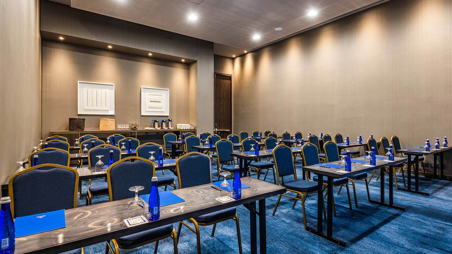 Salón con una decena de mesas rectangulares ubicadas en tres filas horizontales. En cada mesa hay botellas de agua y copas de cristal. Hay 3 sillas por mesa. Al fondo se observa una mesa con unas cafeteras y varios vasos de plástico.