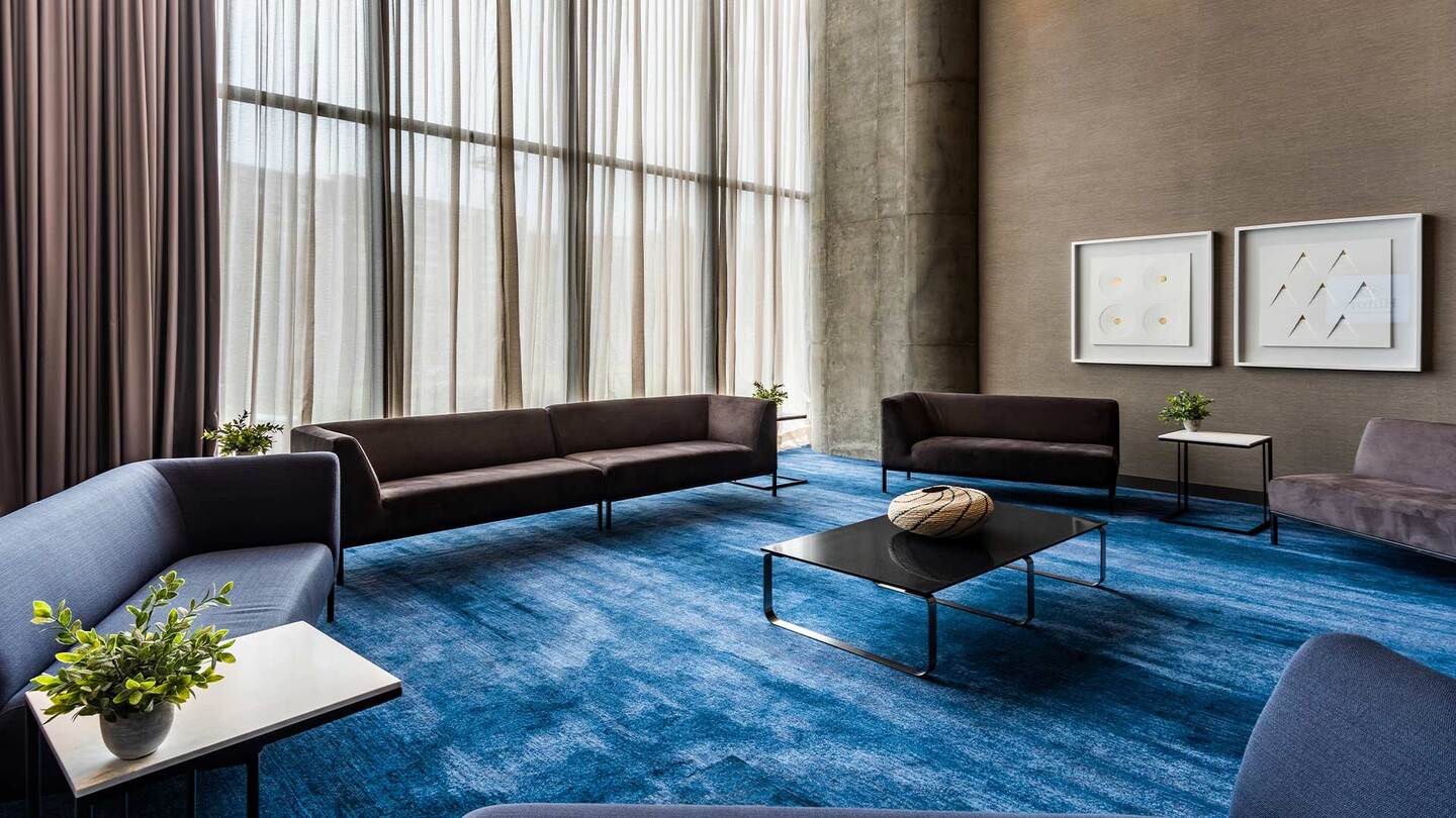 Salón de recepción con por lo menos 5 sofás de color gris y que rodean una mesa de cristal y metal. Hay 4 mesas pequeñas donde sobresale un florero con flores verdes. El piso está cubierto por un tapete azul. Hay un ventanal grande al fondo, por donde entra muy buena luz natural.