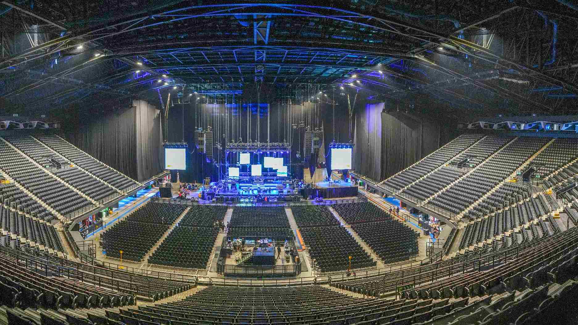  Interior de gran escenario de conciertos vacío y con techo, en el que se observa desde arriba una tarima con pantallas gigantes. Alrededor hay tribunas con miles de sillas.