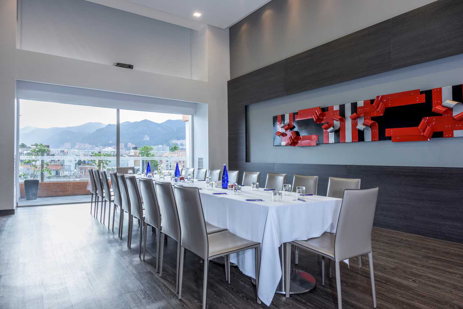 Un salón en un piso alto con una mesa rectangular con mantel blanco, copas y platos. Al fondo hay una ventana donde se observa la ciudad. 