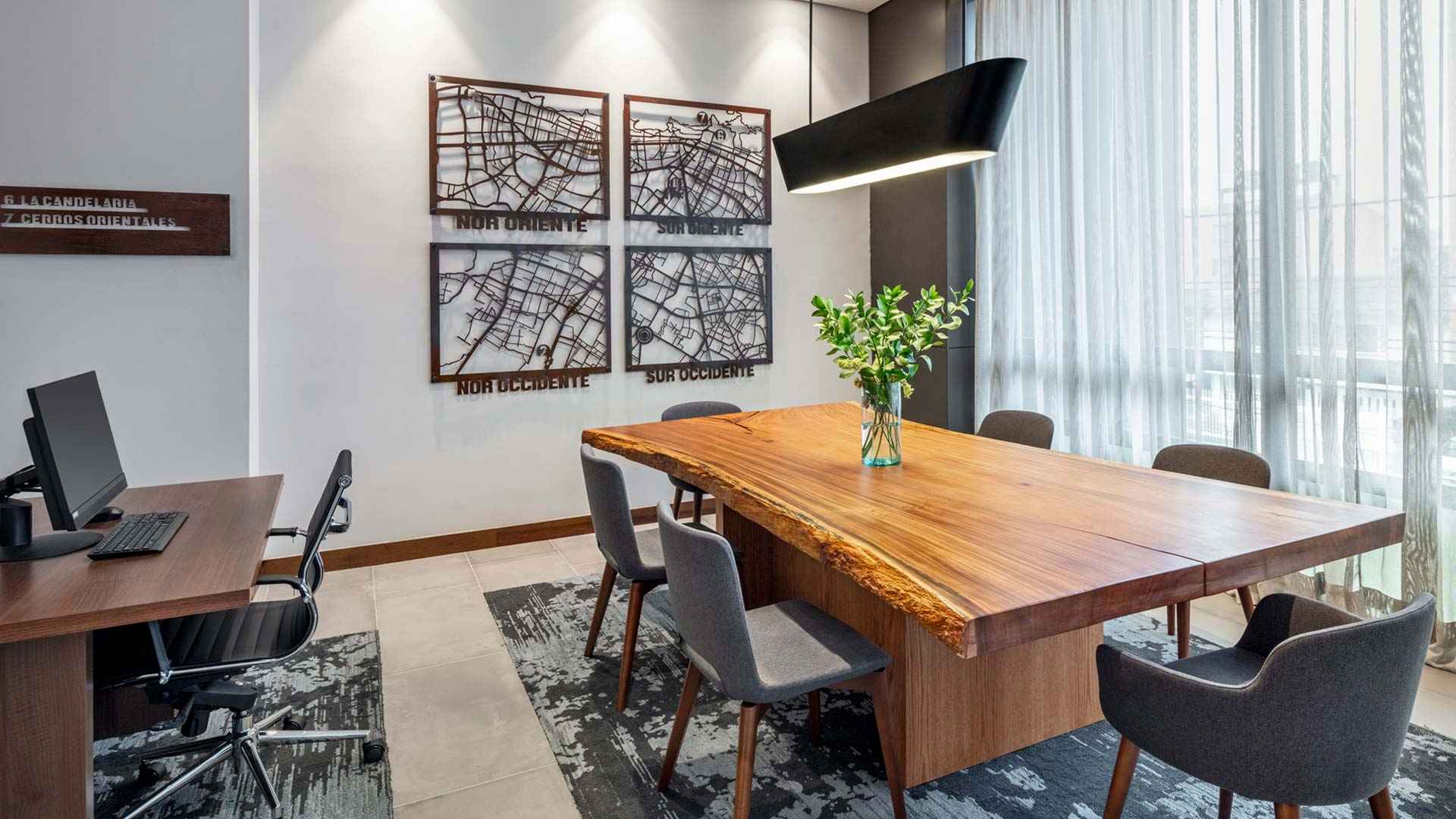 Pequeño salón con una mesa de madera principal y seis sillas alrededor. Una gran lámpara cuelga del techo sobre la mesa. Hay un escritorio con un computador y una silla. En una pared de freente se observan unos mapas de varias zonas de Bogotá.