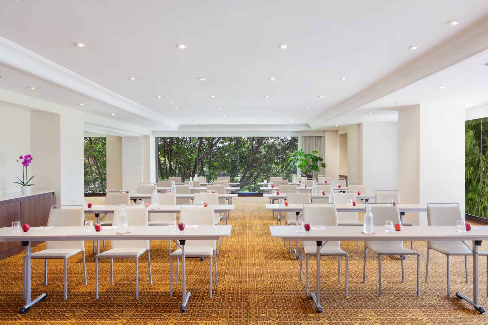 Salón con ventanales a jardines a los lados y al fondo. Hay dos filas con 10 de mesas, organizadas de forma vertical. hay un lindo tapete color caoba y una matera con una orquídea.