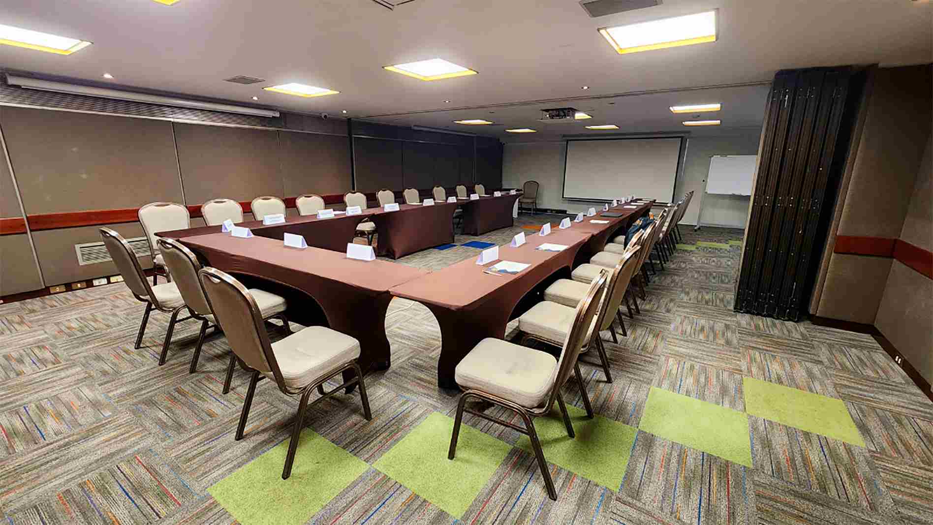 Salon con una gran mesa en U, decorada con manteles de color marrón. Alrededor de la mesa hay varias sillas blancas. Al frente hay una pantalla de videobeam.