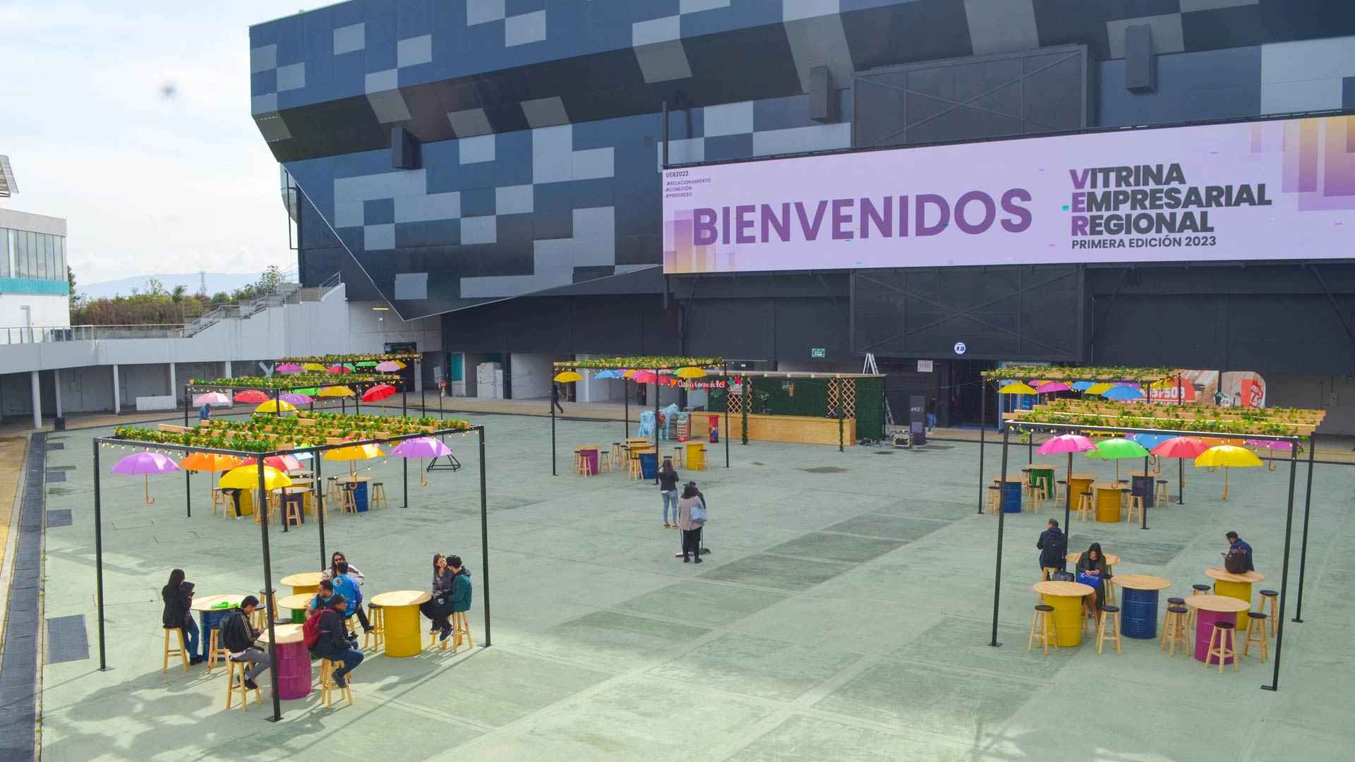 Outdoor plaza during the day with several cubicles with tables surrounded by people sitting on chairs. In the background, a tall concrete structure features a sign that reads “Welcome, regional business showcase.”
