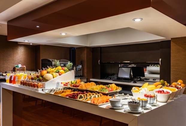 Salón en el que hay una estantería amplia sobre la que hay trozos de frutas y botellas de jugo. Al fondo hay un computador y una cafetera.