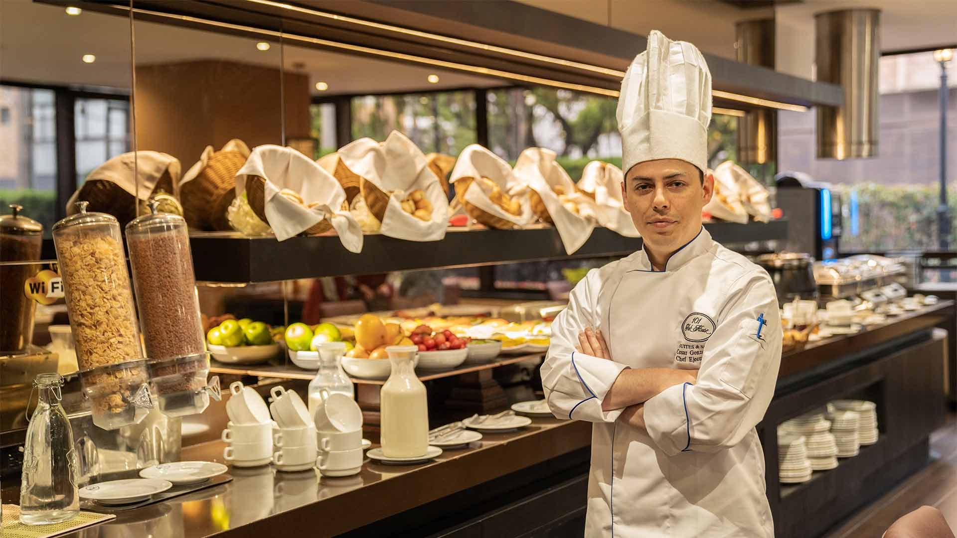  Hombre joven chef al frente de una estantería de vidrio y madera en la que hay canastas con panes, badejas con frutas y recipentes con líquidos.