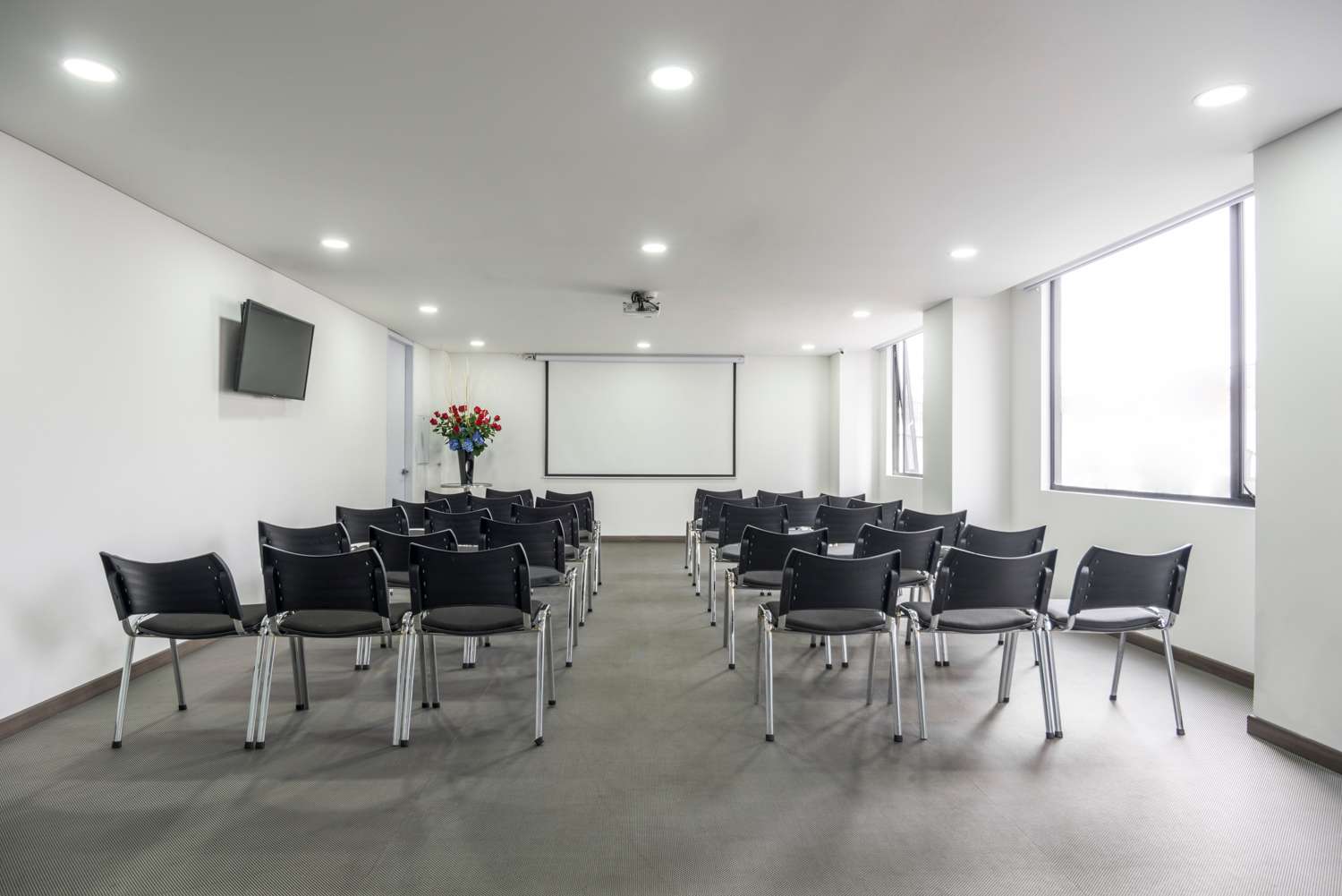 Salón con paredes y techo blanco, y el piso con baldosa de color negro. Hay seis filas de sillas de plástico en forma vertical mirando a un tablero. En una pared hay una pantalla de televisión. En la otra hay ventanas.