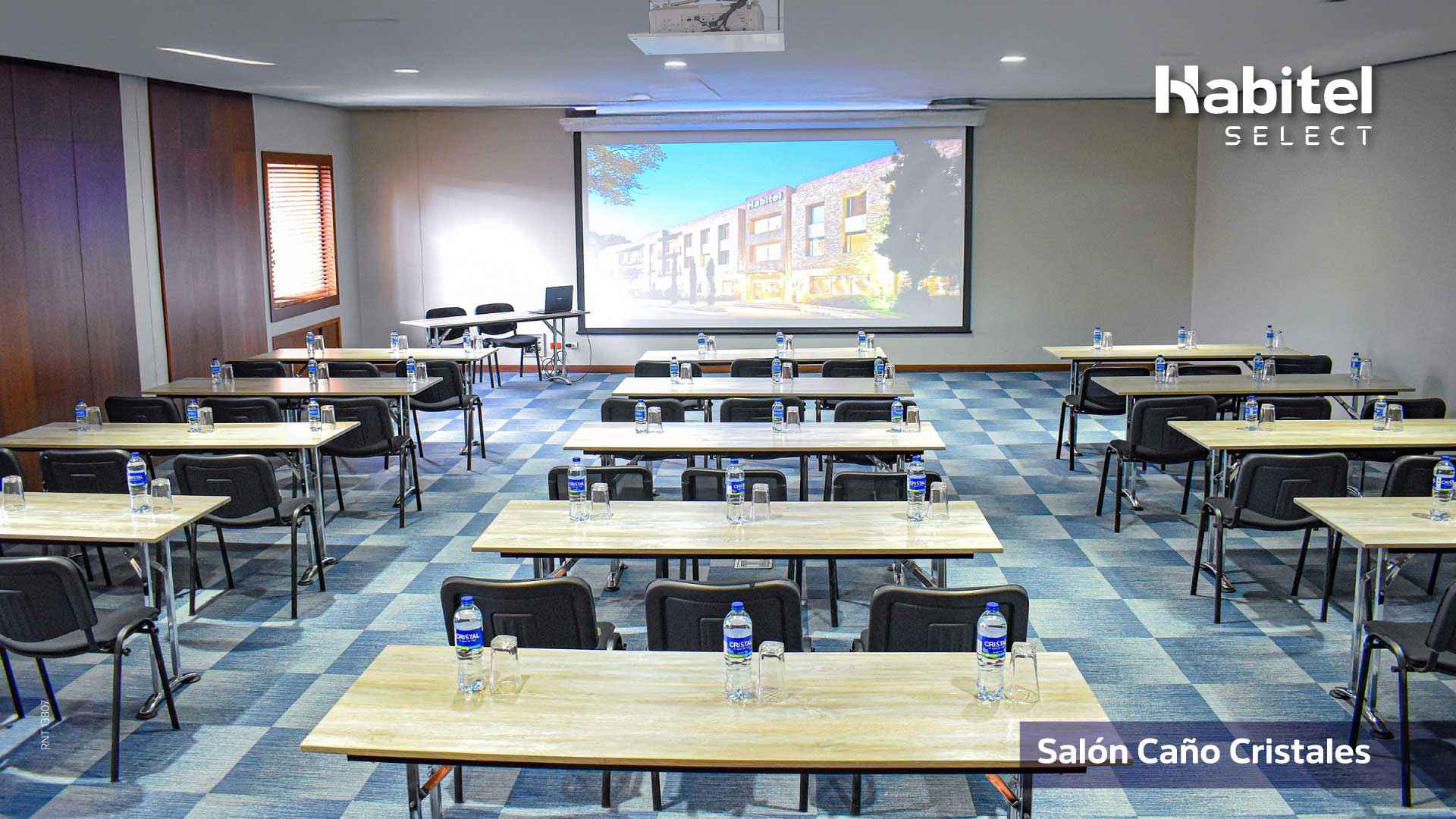 Salón dispuesto para conferencia con cerca de 15 mesas de a tres sillas cada una. En cada mesa hay botellas con agua y vasos de vidrio. Al frente de las mesas hay una pantalla de videobeam.