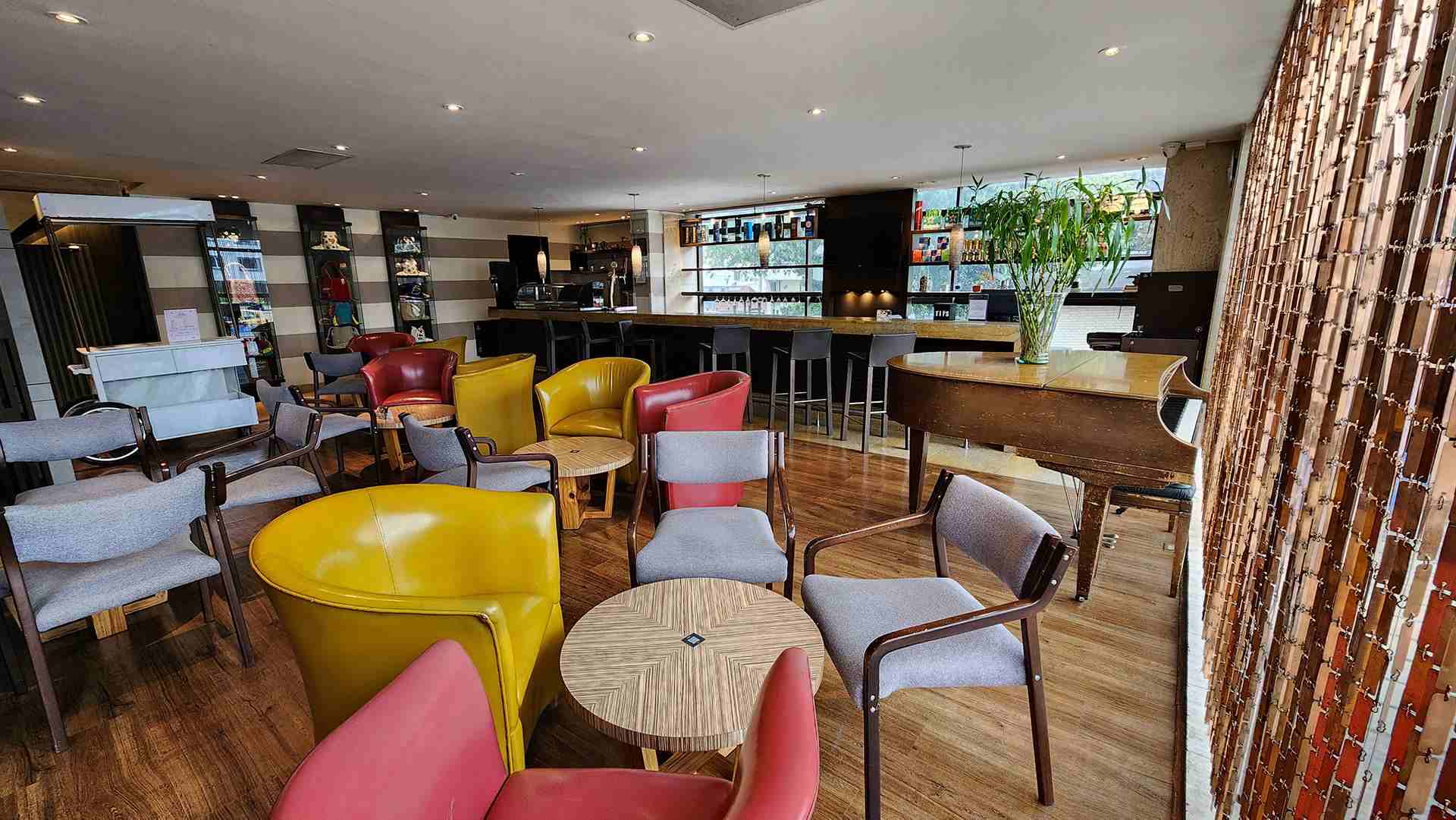 Salón pequeño en el que hay varias sillas y poltronas rodeando mesas redondas. Hay un piano de cola y una barra estilo bar.