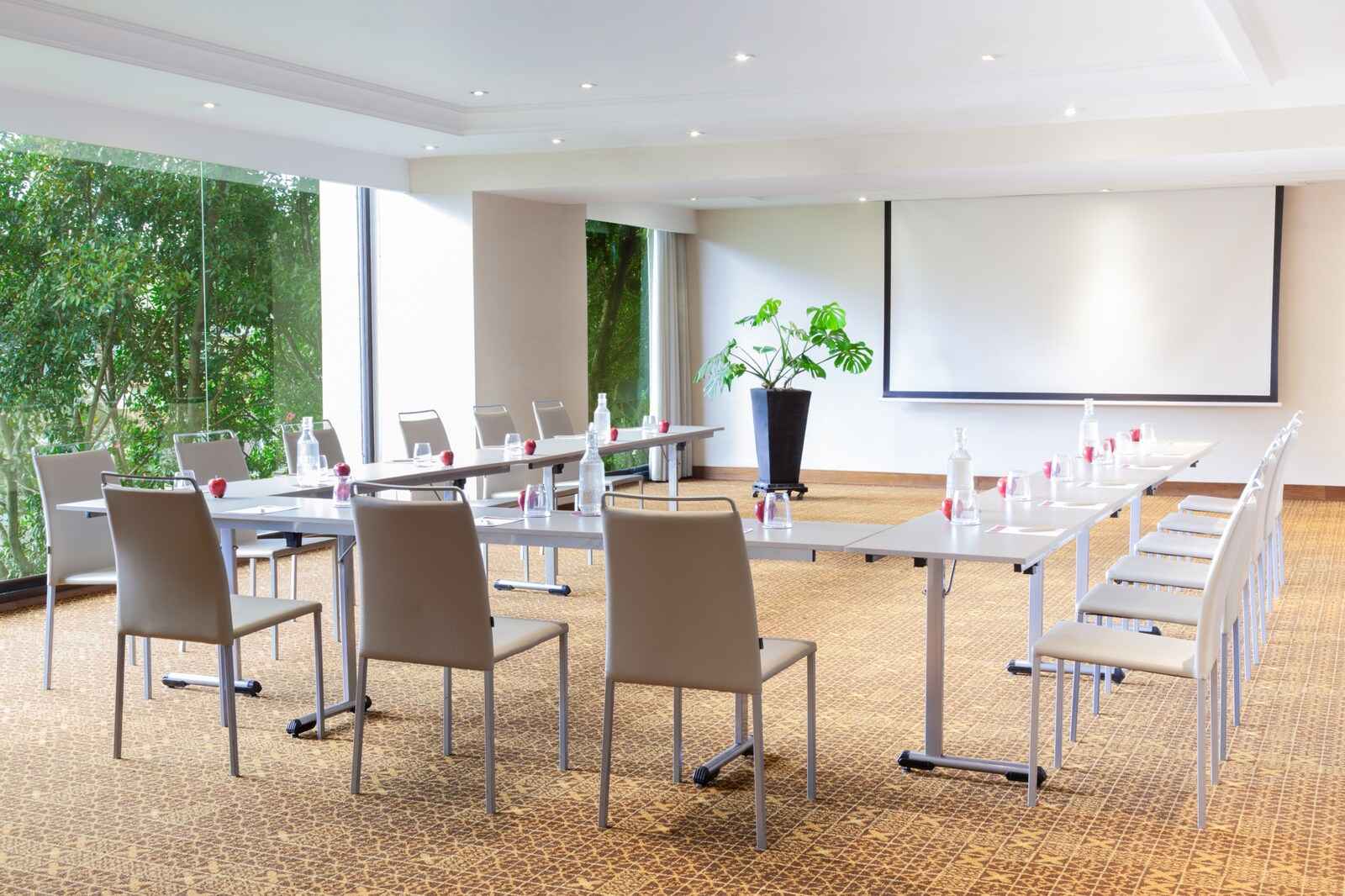  Salón con ventanales a jardines a los lados. Hay varias mesas organizadas en forma de U. En cada mesa hay manzanas y botellas de agua. Al frente hay una pantalla de videobeam y una matera con una planta.