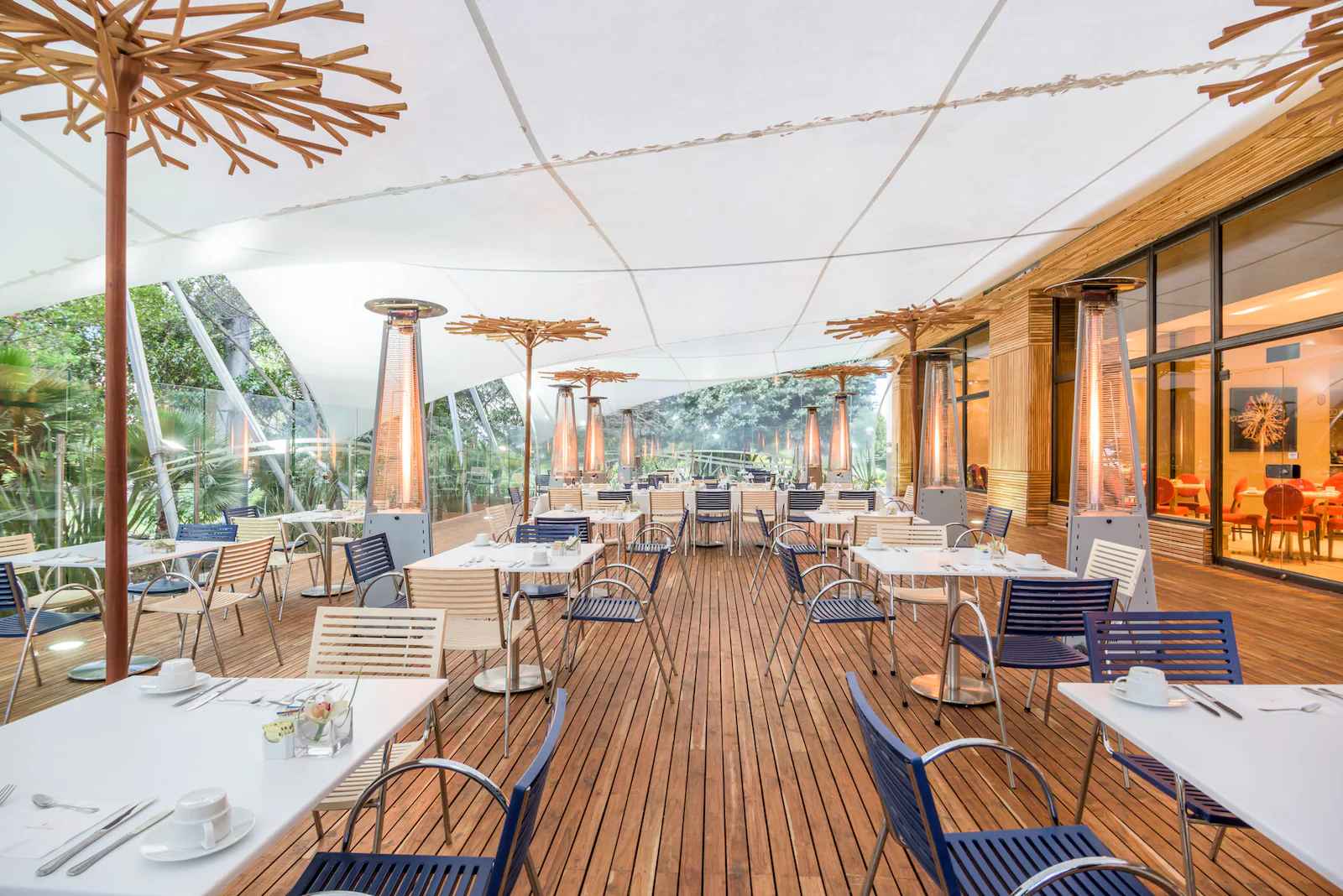 Terraza interior decorada con piso en madera y un techo en lona blanca. Hay varias mesas con sillas rodeadas por varios calentadores de ambiente en forma de triángulo.