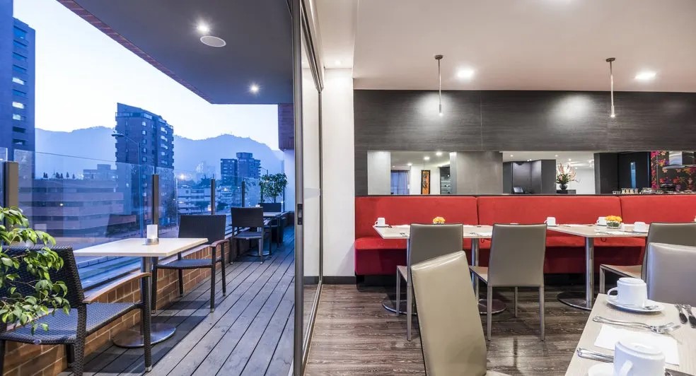 Salón en un piso alto en el que se observa por un lado una terraza exterior con mesas pequeñas y sillas, y por el otro un salón con mesas y sillas.