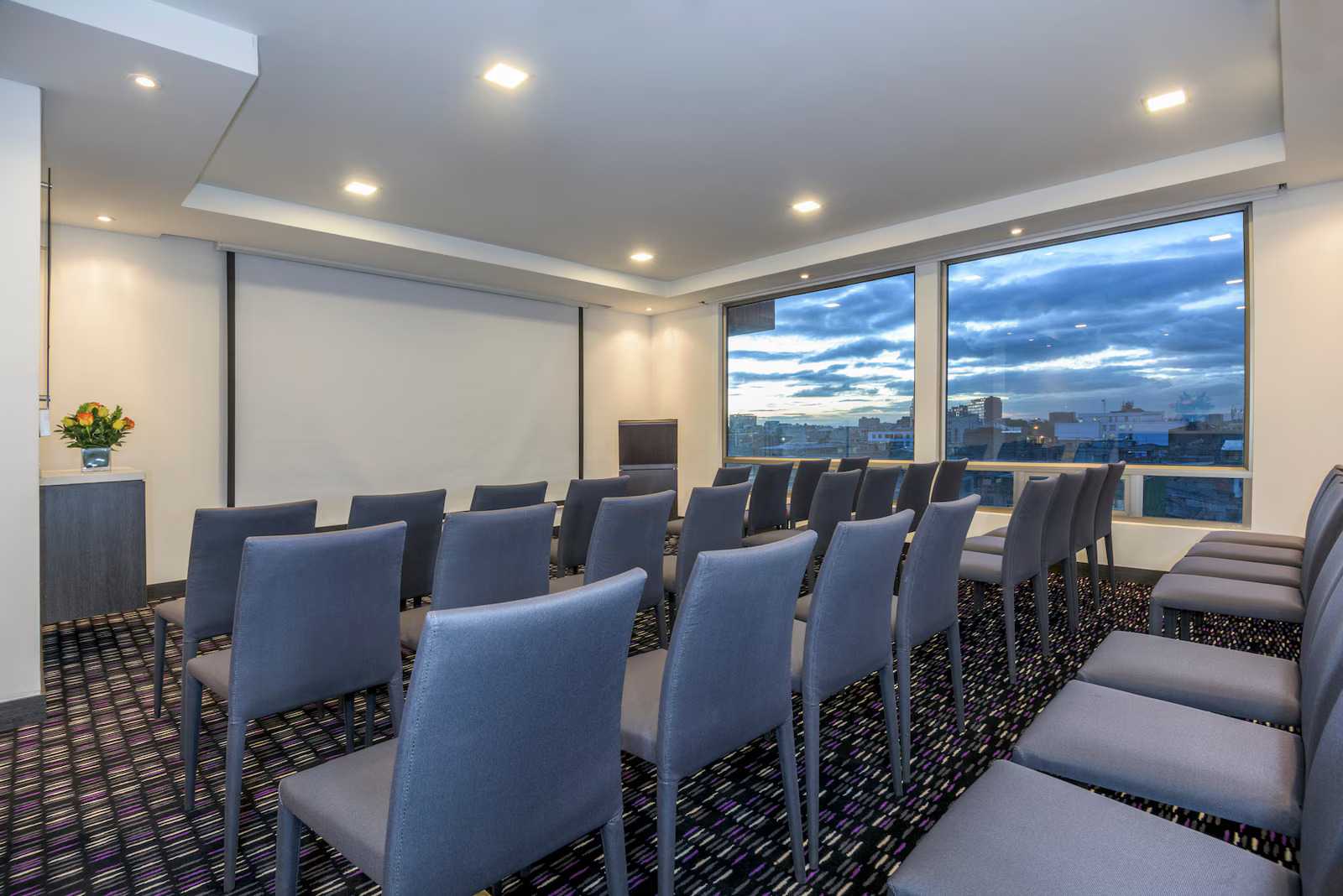 Pequeño salón de reunión en un piso alto donde hay algunas sillas orientadas hacia una pantalla gigante. Hay un ventanal donde se ve la ciudad al atardecer.