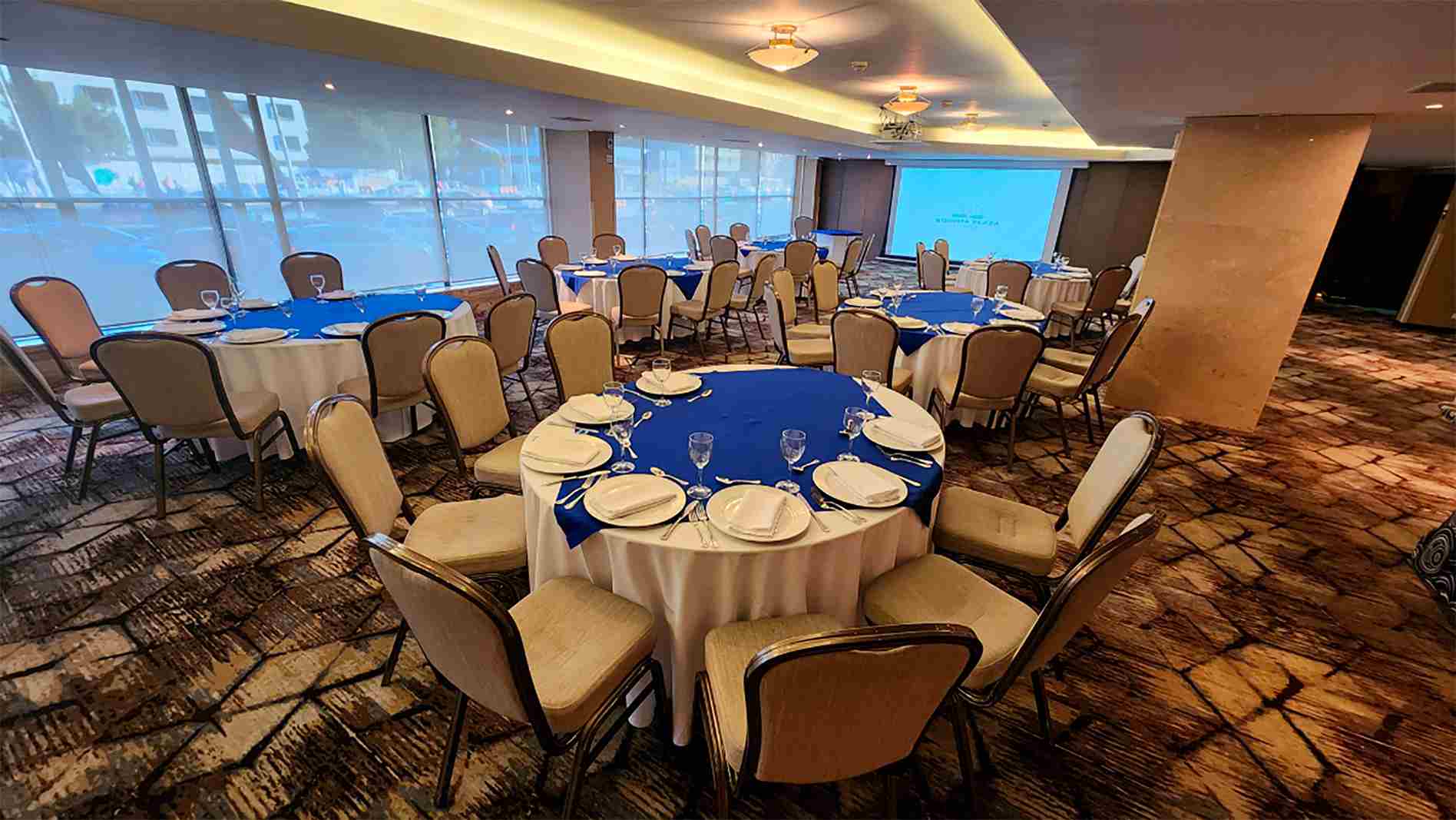 Salón con mesas redondas y sillas alrededor. Las mesas están decoradas con manteles, cubiertos y copas de cristal. A un lado hay un gran ventanal y al fondo una pantalla de videobeam.