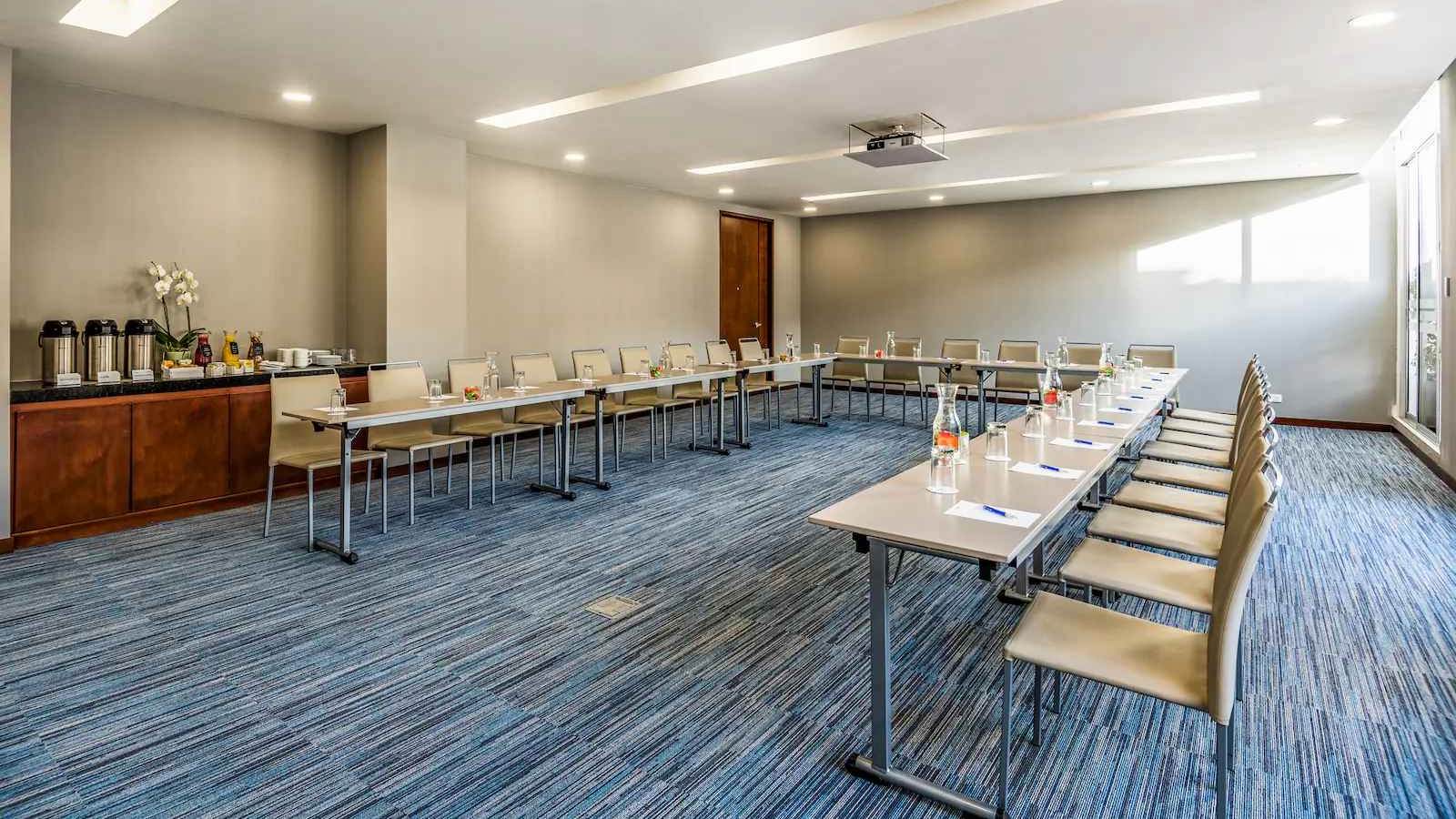  Salón amplio con una mesa organizada en forma de U y varias sillas alrededor. Hay una mesa al lado de una pared con varias cafeteras.