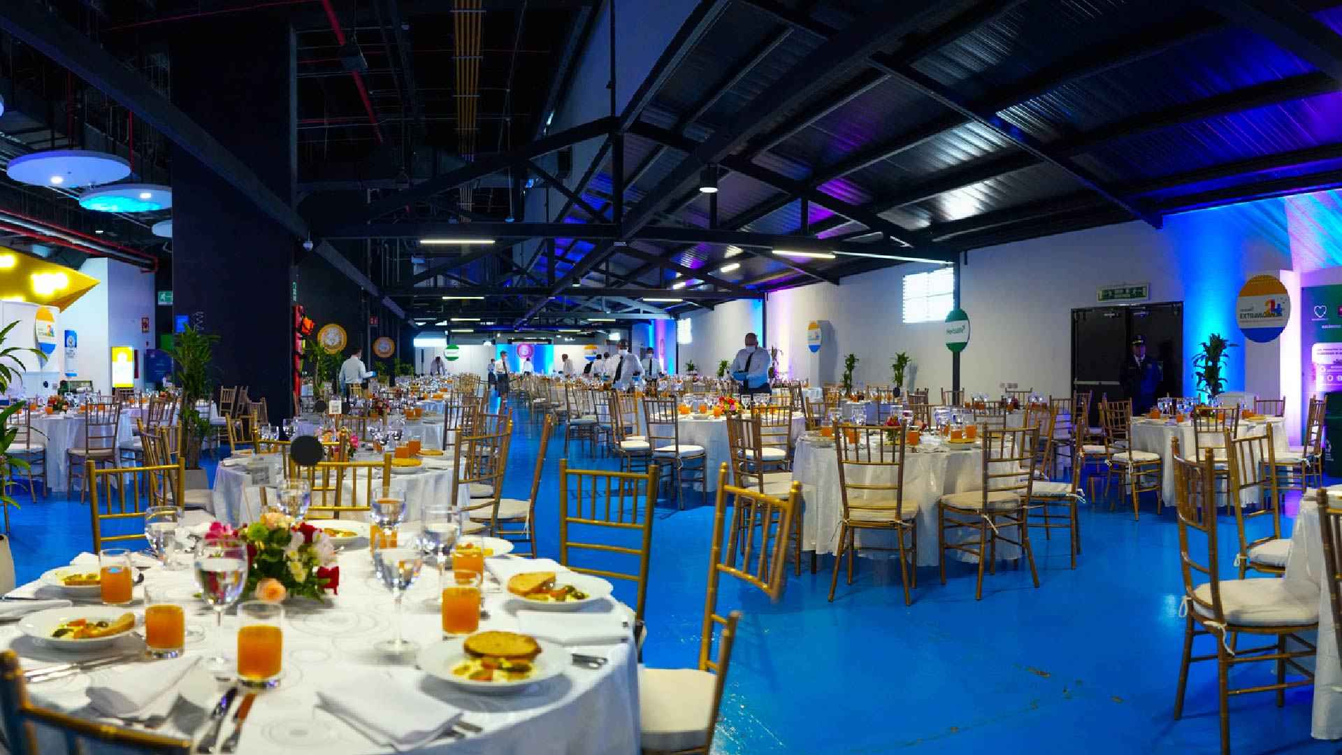 Interior de un salón decorado para un evento social. Se ven mesas redondas con copas de cristal, platos con alimentos, y cubiertos. Hay meseros alrededor.