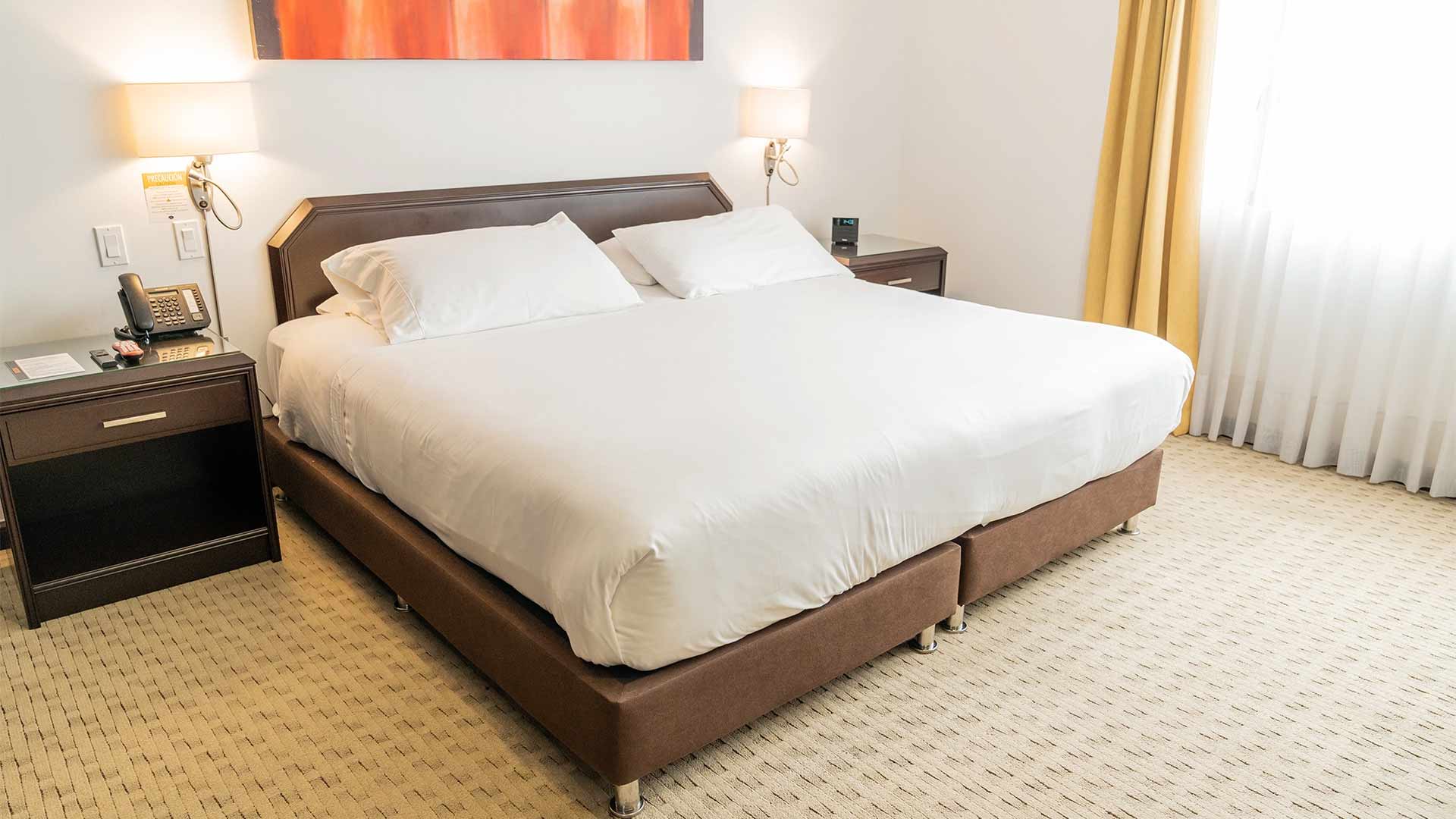 Habitación con una cama grande con dos mesas de noche, una a cada lado. En una mesa hay un reloj despertador y en la otra un teléfono. Hay dos lámparas a cada lado de la cama.