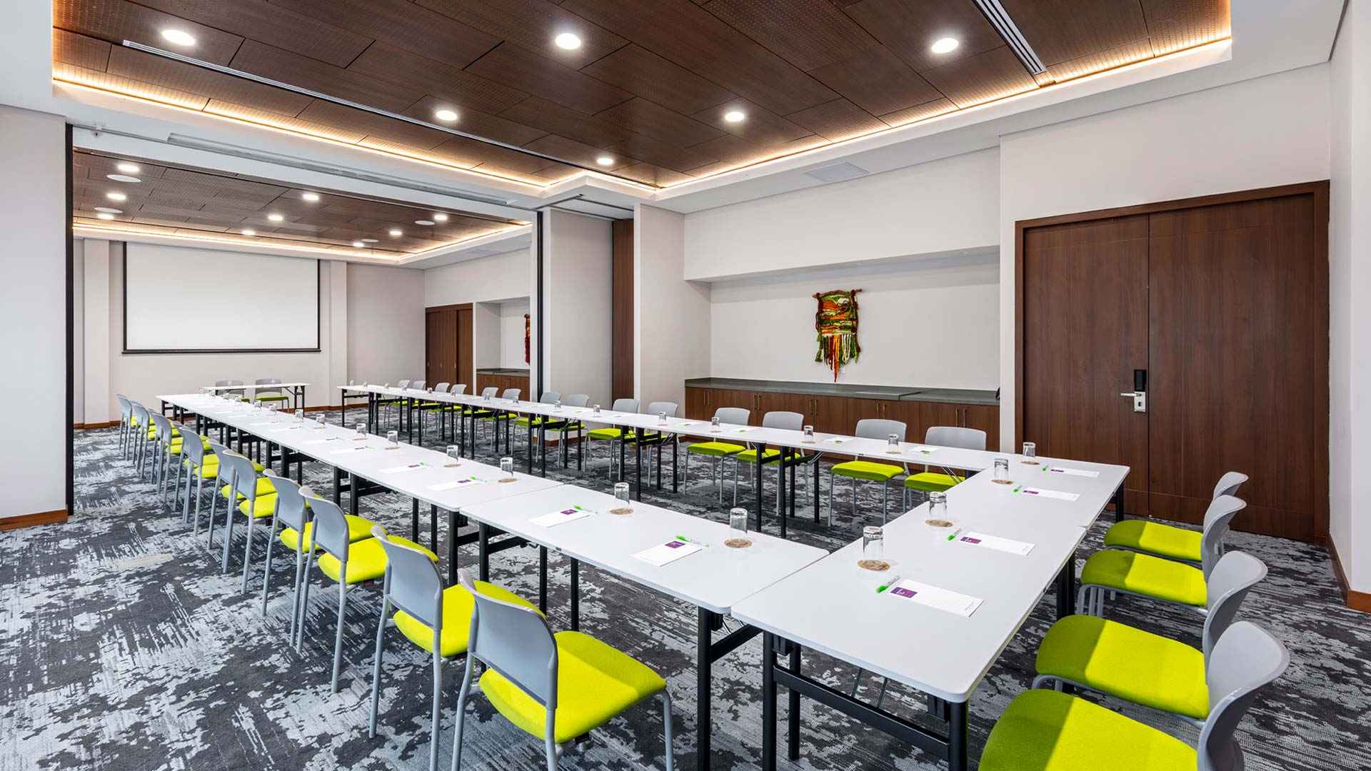 Gran salón con varias mesas blancas formando una U. Hay varias sillas de color blanco y amarillo alrededor. Al fondo hay una pantalla de viodeobeam.