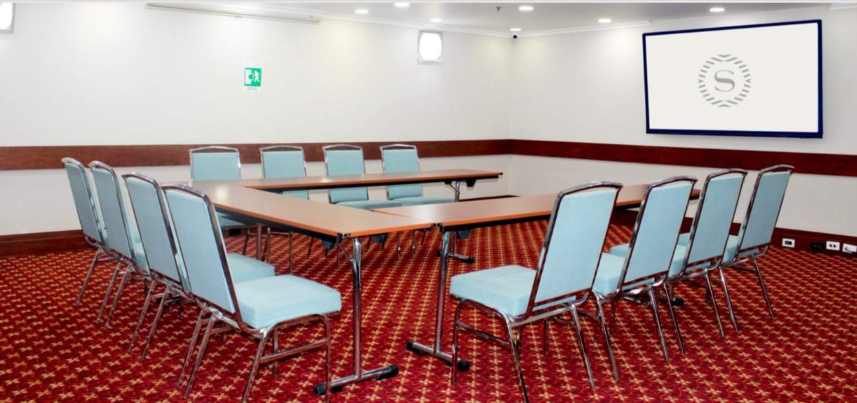 Salón pequeño entapetado con mesas organizadas en forma de U. Hay doce sillas rodeando la mesa y una pantalla de televisión al frente.