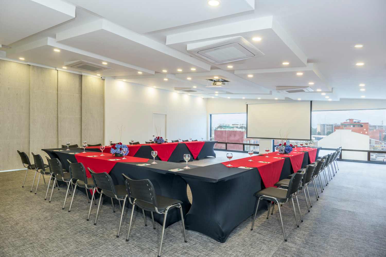 Gran salón en forma de U con las mesas forradas en tela negra con manteles rojos. Sobre las mesas hay copas de vidrio. Al frente del salón hay un ventanal gigantesco.