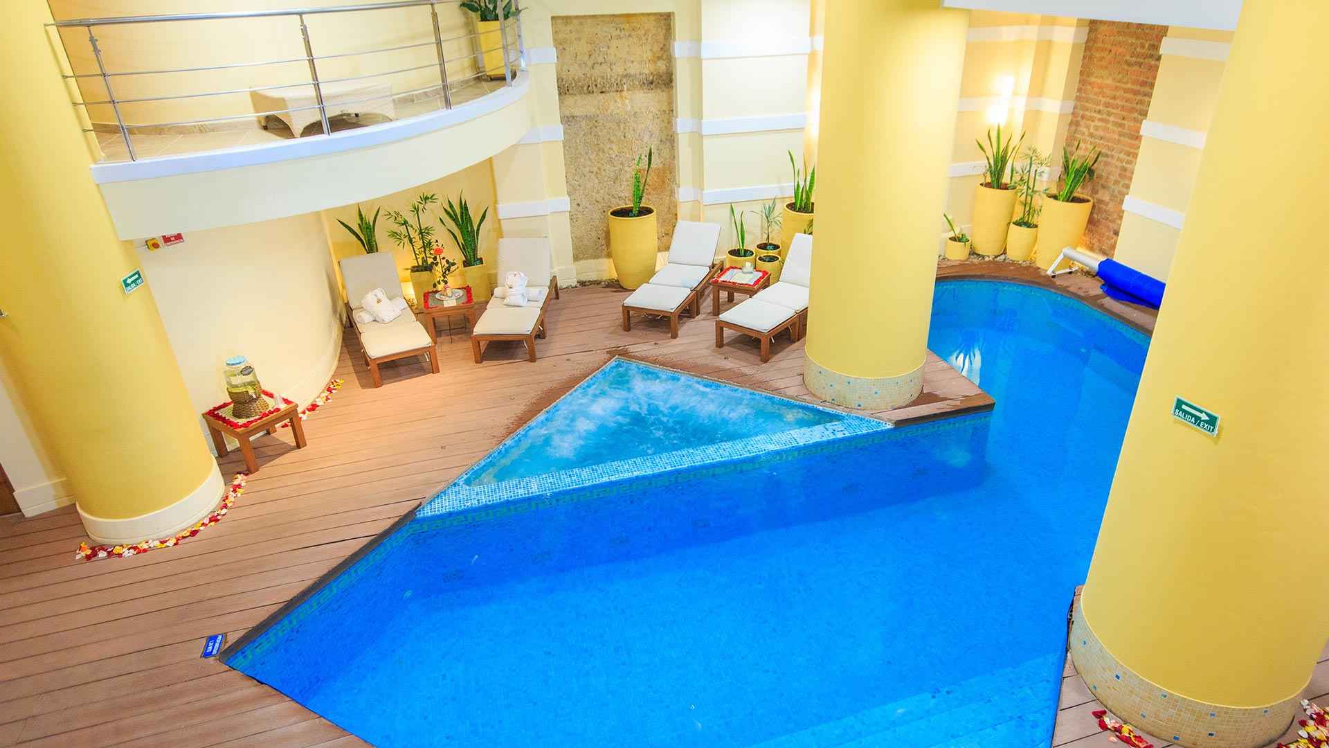 Salón interior con una gran piscina de color azul. Destacan tres columnas gigantes y redondas. Alrededor de la piscina hay tres sillas reclinables.