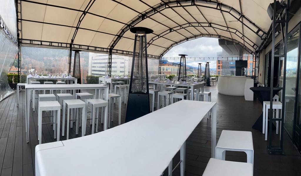 Terraza en piso alto, cubierta por un techo en lona blanca. Hay varias mesas y sillas blancas  y varios calefactores en forma de pirámide. Al fondo se observa la ciudad.