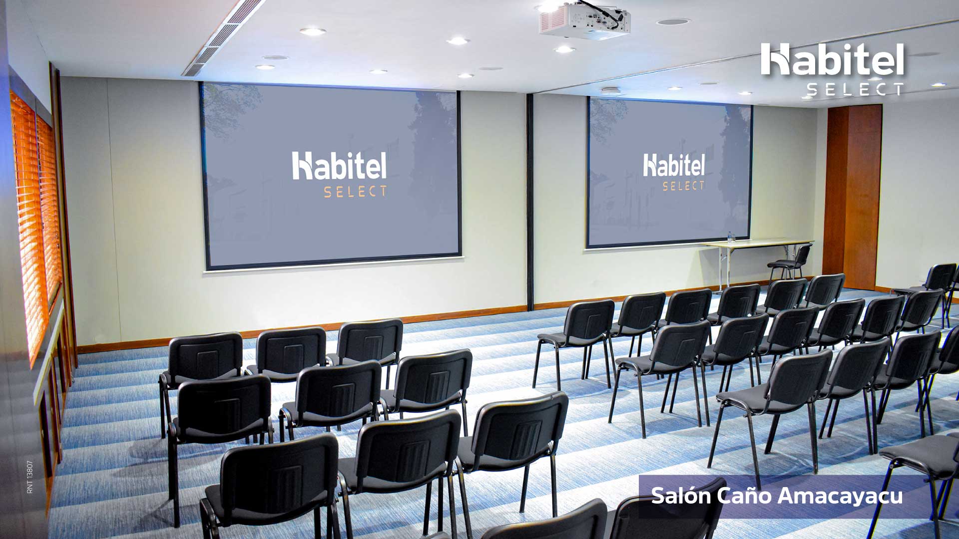  Salón de reuniones en el que se ven casi 20 sillas metálicas negras, mirando a dos pantallas de videobeam al frente.