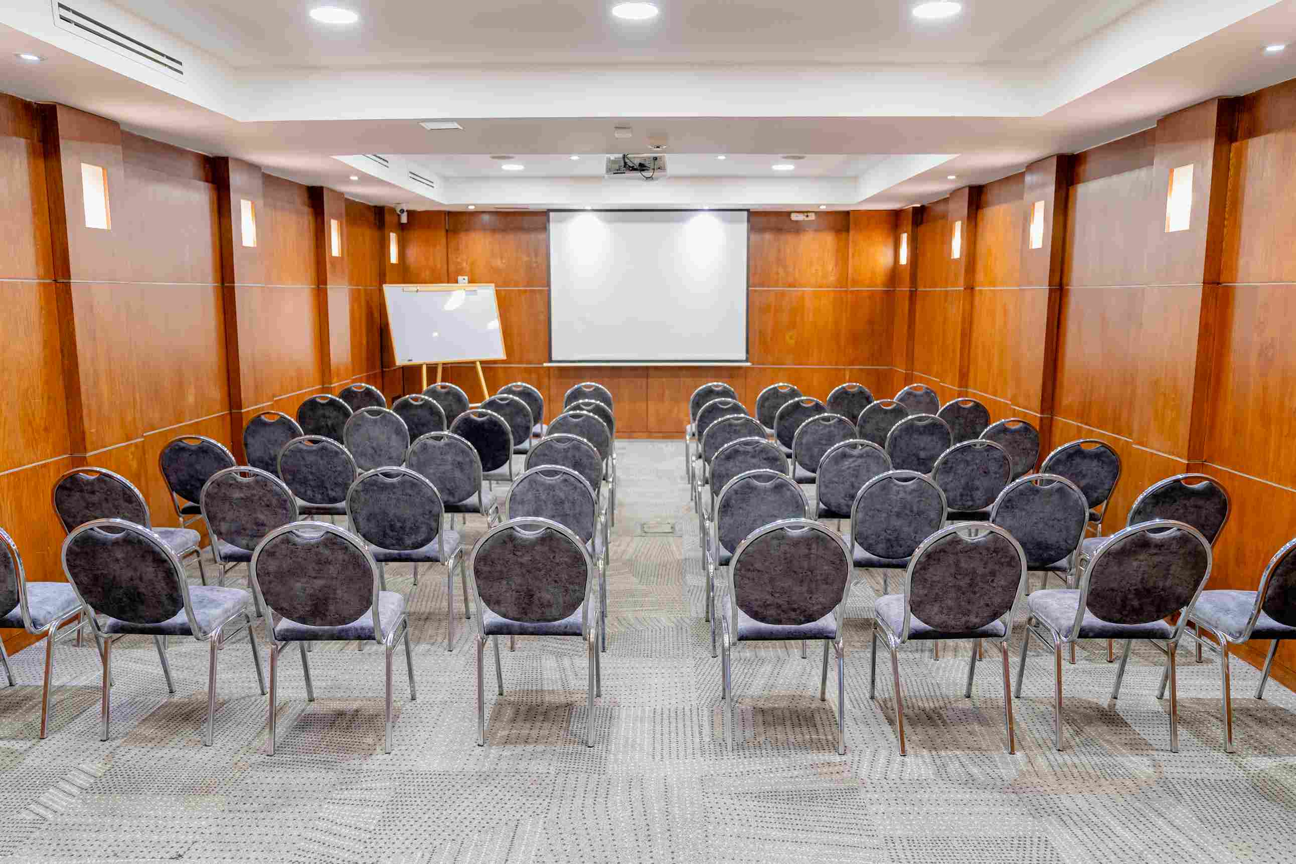 Salón pequeño con Salón con paredes de madera y por lo menos 50 sillas grises. Al frente hay una pantalla de videobeam.