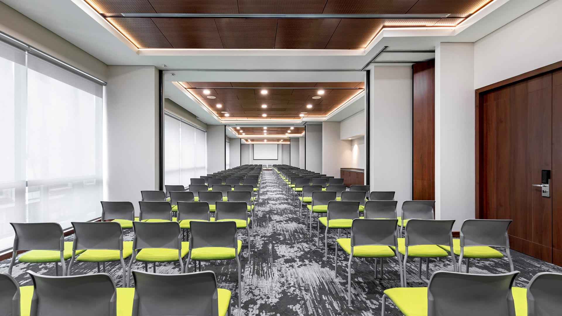Gran salon rectangular con un centenar de sillas. Hay muchas ventanas a un costado. Al fondo hay una pantalla de viodeobeam.