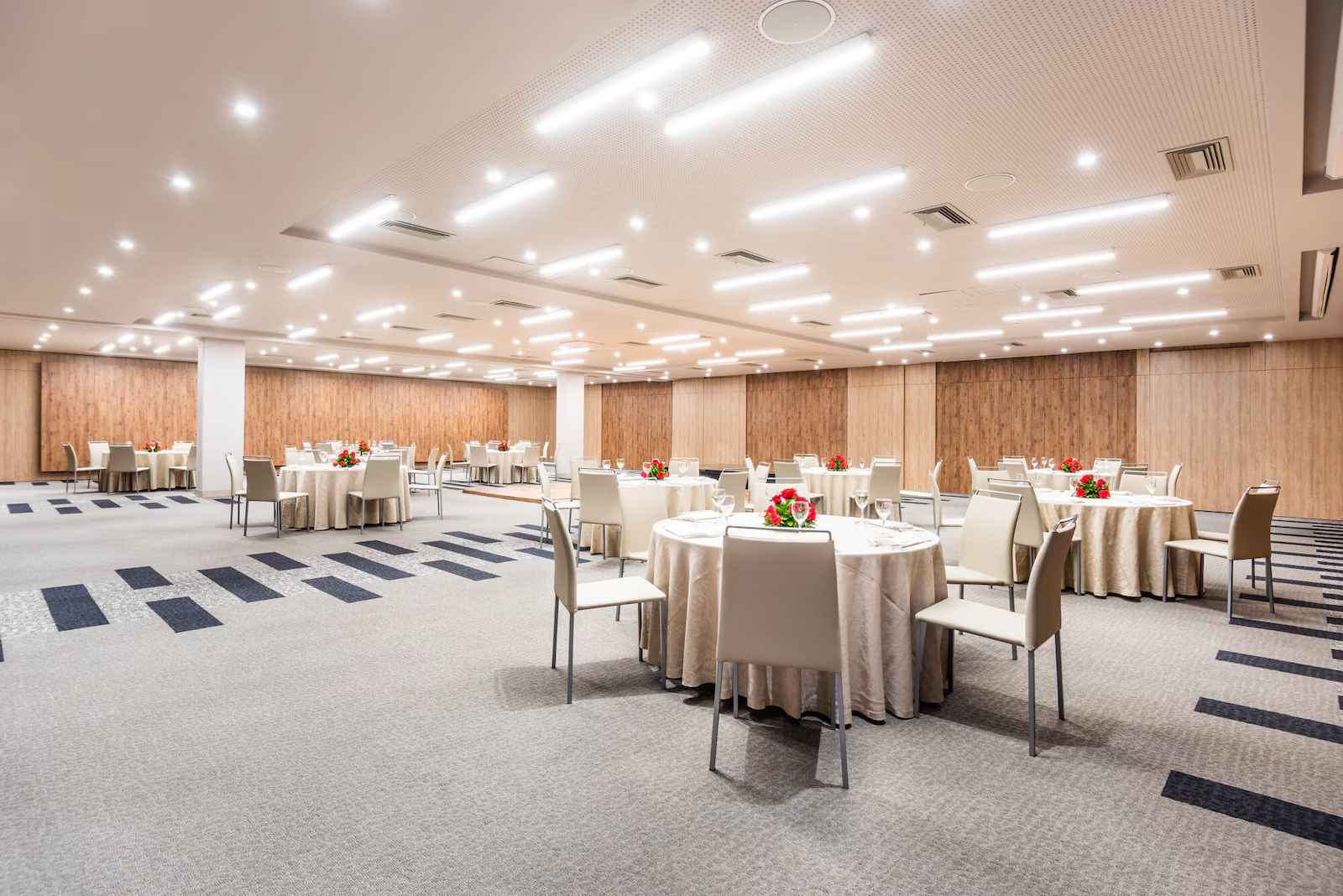  Gran salón acondicionado con mesas redondas y con manteles, dispuestas para un evento social. Hay arreglos florales en casa mesa.