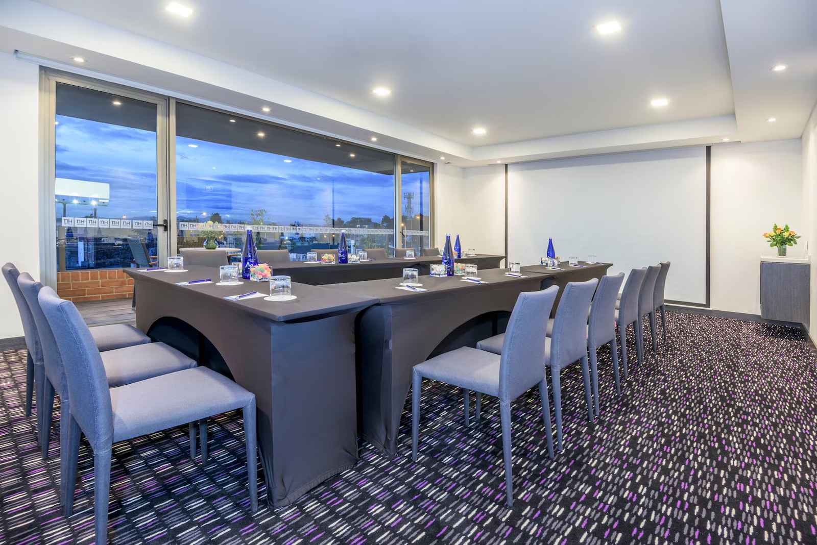 Un salón en un piso alto con una mesa rectangular con mantel gris y vasos de vidrio. Hay una pantalla gigante y al fondo hay una ventana donde se observa la ciudad al atardecer. 