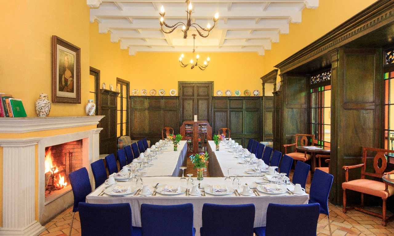 Salón antiguo con una mesa en U, vestida con mantel blanco y rodeada de sillas con tapiz de color azul. A un lado hay una chimenea encendida. Hay una colección de platos colgados en una pared.