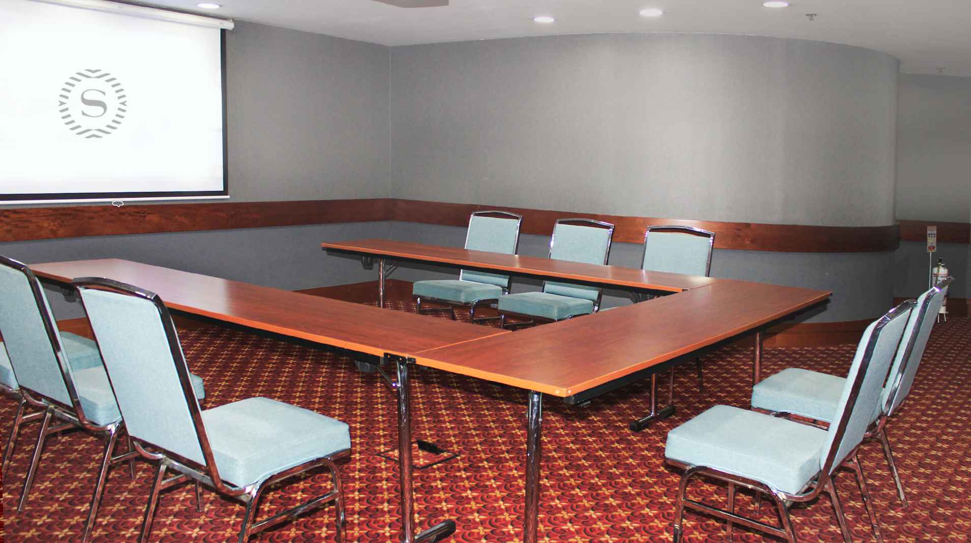 Un salón con una mesa en forma de U rodeada de por lo menos 6 sillas. hay una pantalla de videobeam al frente en la que se proyecta una letra S.
