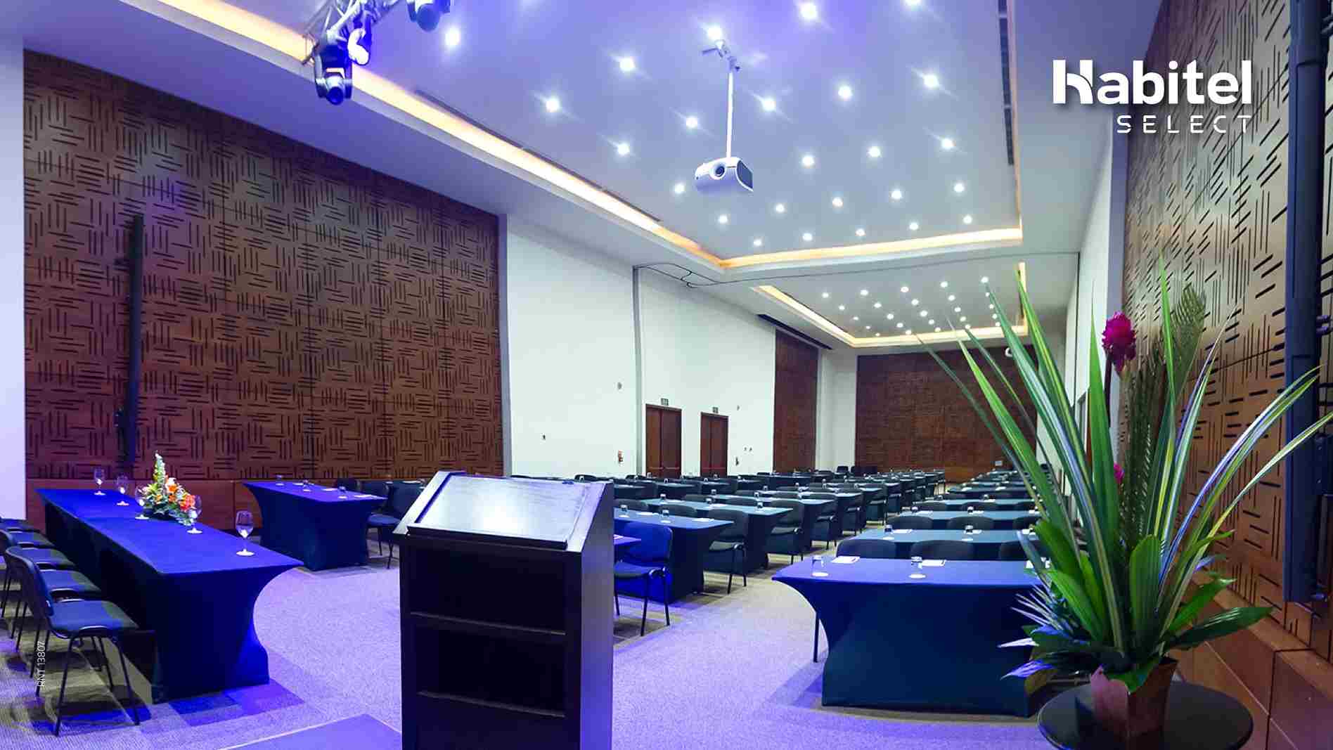 Salón rectangular dispuesto para conferencia. Hay varias mesas decoradas con manteles azules. Hay una mesa principal y un atrio.