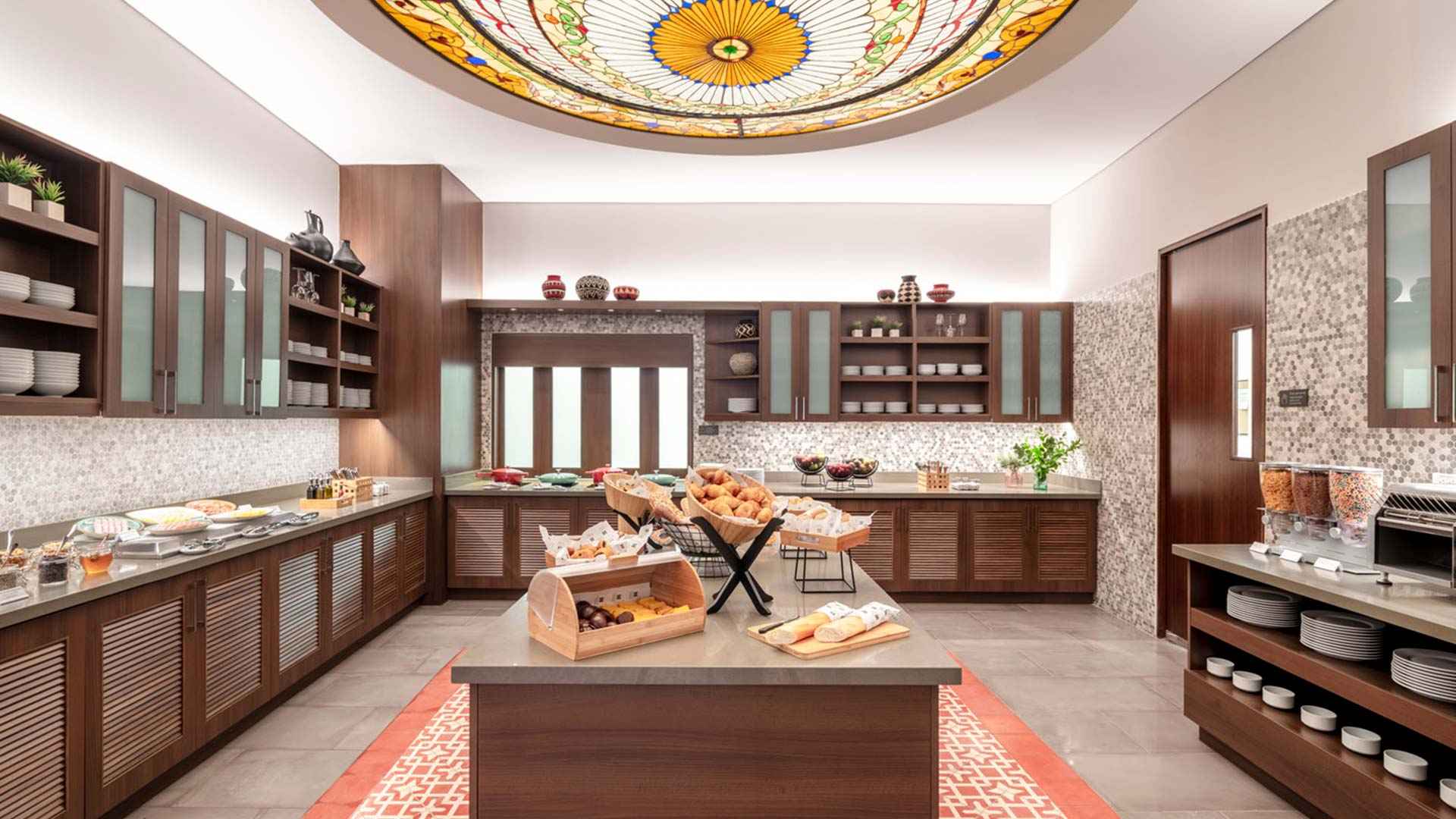 Cocina elegante decorada con estates de madera. Sobre dos mesones largos hay canastas con pan, frutas y algunos recipientes con líquidos. Hay otro mesón lleno de platos y pocillos. En el centro hay un mesón con varias canastas y recipientes con pan. Del techo cuelga una lámpara gigantesca y con estilo vitral.