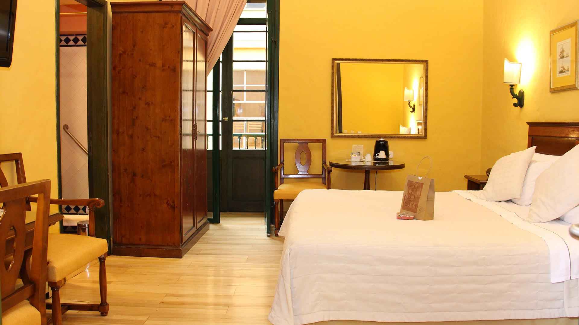 Habitación de color amarillo con una cama grande, un espejo, una silla, una lámpara en la pared y un closet, todo de estilo antiguo como de los años 40.