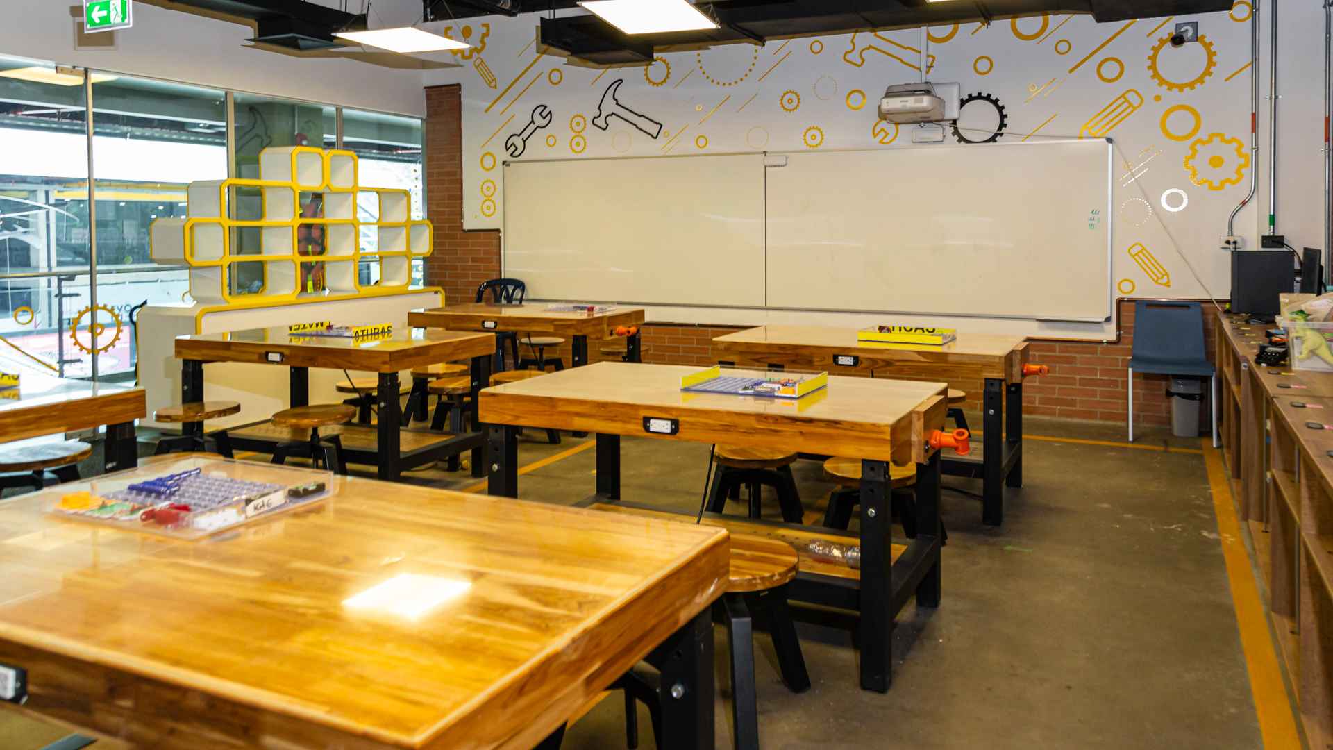 Salón pequeño con 6 mesas de madera gruesa color dorado. Hay alrededor varios butacos de madera del mismo color. A espalda de las mesas en una pared hay dos tableros acrílicos. Un videobeam cuelga del techo.