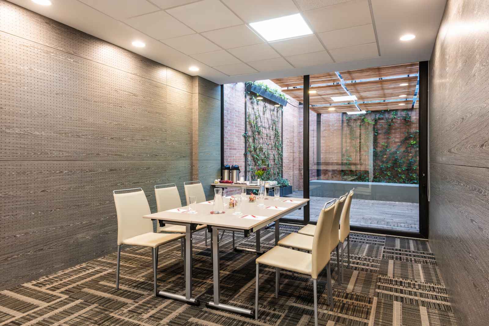 Salón muy pequeño en el que sobresale una mesa con 6 sillas alrededor. A un costado hay una mesa con dos cafeteras y varios pocillos. Al frente hay un ventanal hacia un patio interior. 