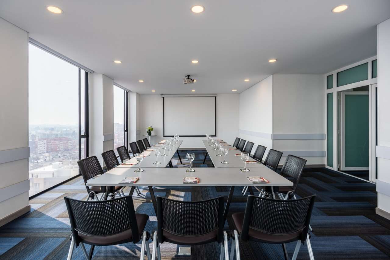 Salón pequeño y rectangular con mesas formado una U. Alrededor hay varias sillas. A un costado hay dos ventanales grandes.