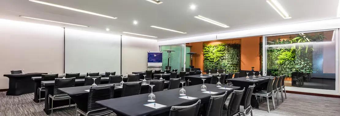 Gran salón en el que destacan varias mesas con manteles negros y rodeadas de sillas de igual color. En cada mesa hay copas de cristal. Al frente de las mesas hay una pantalla de videobeam. 