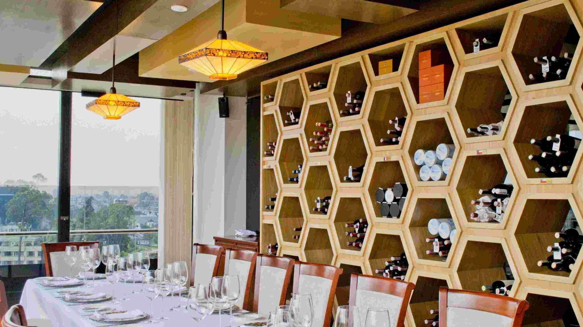 Interior of a room with an elegant, well-stocked wine cellar behind a long table decorated with white tablecloths, crystal glasses, and plates.
