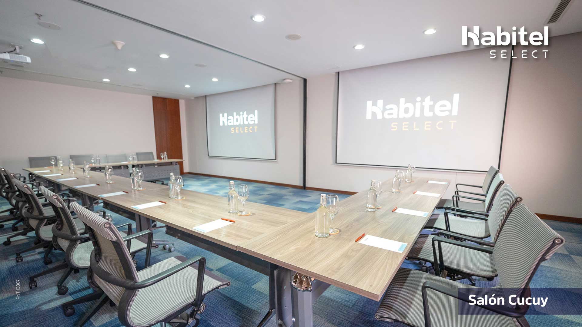 Salón con una gran mesa en forma de U. Alrededor de la mesa hay sillas y una pantalla de videobeam al frente.